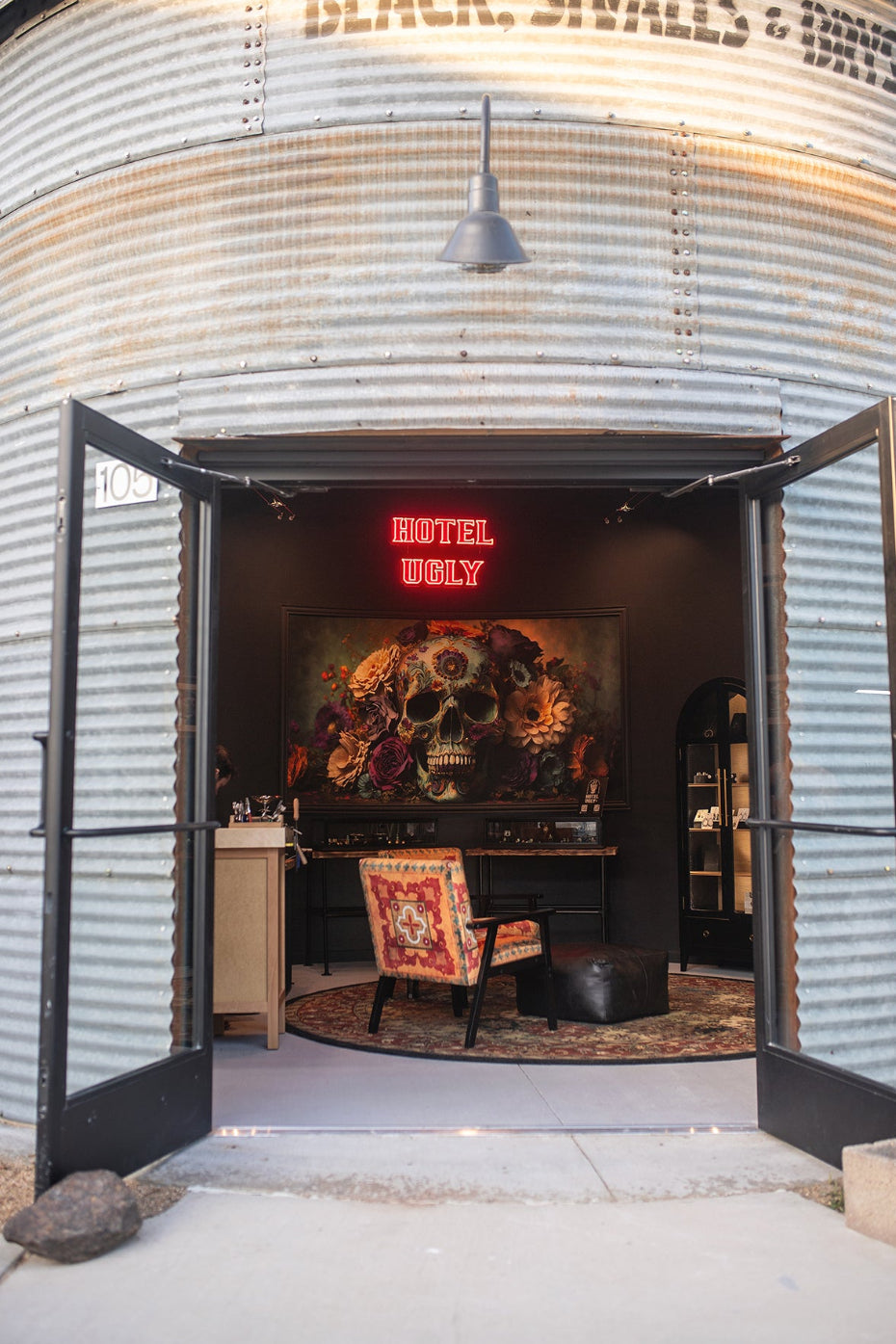The entrance to Hotel Ugly, featuring open double doors set into a corrugated metal building. Inside, a gothic-themed room is visible with a large skull painting, a red neon sign, and a patterned armchair.